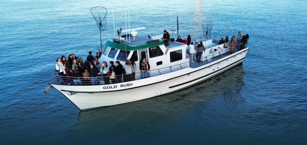 A group of people stand and sit on the deck of a white boat named "Gold Rush," a private charter floating on calm blue water, perfect for Alcatraz Island Cruises and Fisherman's Wharf departures.