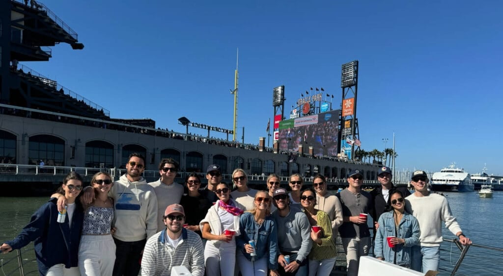 A group of people pose together on a boat during one of the San Francisco Bay Cruises, with a stadium by the waterfront, clear blue sky, and boats in the background.