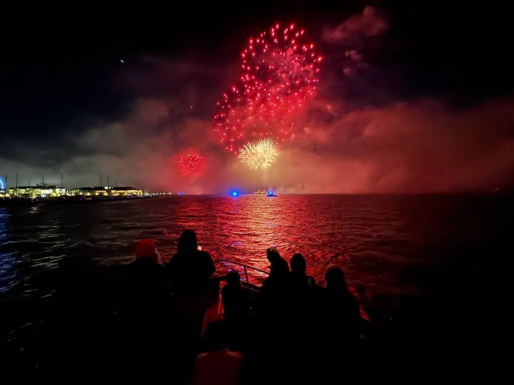 A group of people on a boat watch red and yellow fireworks light up the night sky over the water near a city shoreline during one of the popular Golden Gate Bridge Tours.
