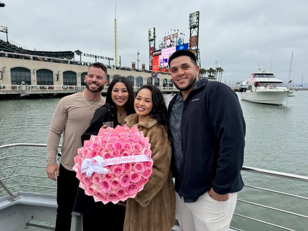 Four people stand on a boat docked in a marina, with one holding a large bouquet of pink roses; a stadium and yacht are visible in the background, capturing the charm of San Francisco Bay Cruises from Fisherman's Wharf Departures.
