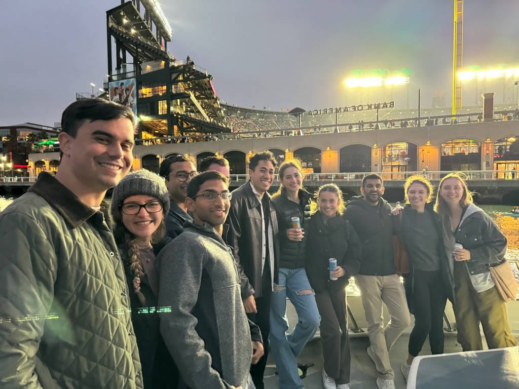 A group of people stand together smiling for a photo outdoors near a stadium at dusk, with stadium lights and seating visible in the background—reminiscent of friends gathering before San Francisco Bay Cruises or Private Charters.