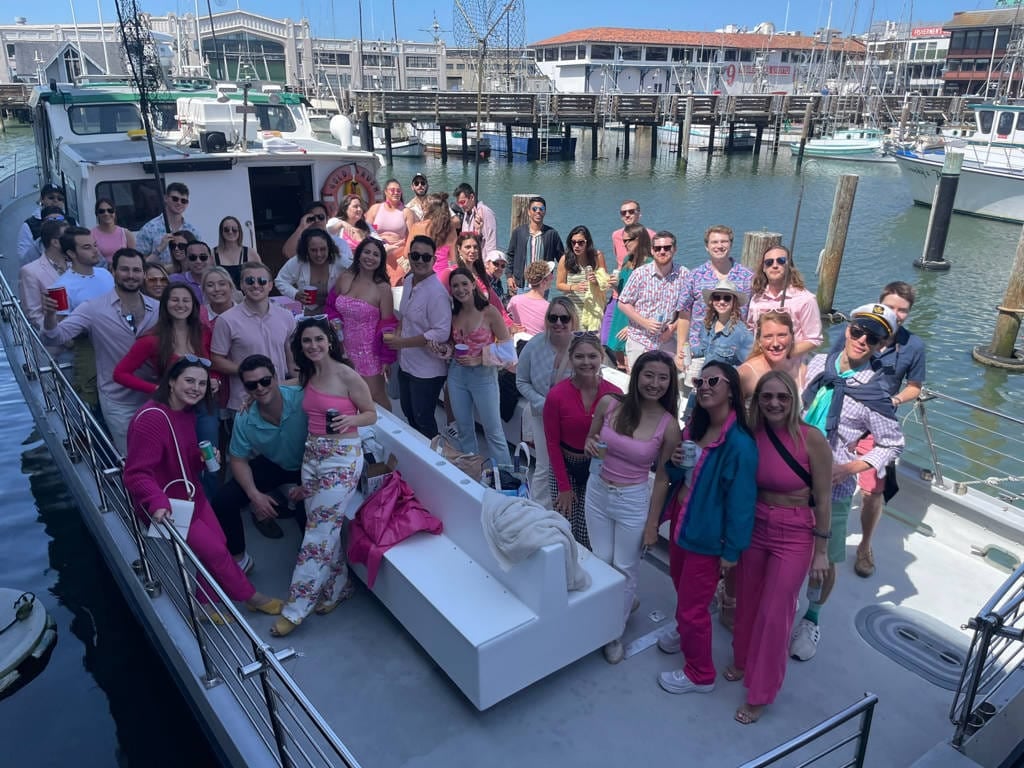 A large group of people dressed in colorful and pink-themed clothing pose together on a boat docked at a marina on a sunny day, ready for one of the Fisherman's Wharf Departures or Private Charters.