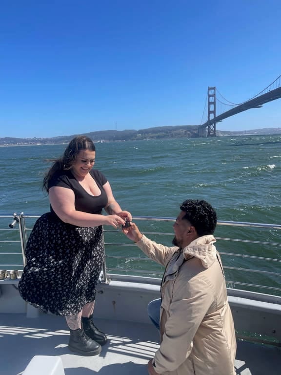 A man kneels and proposes to a woman on a boat with the Golden Gate Bridge in the background, likely during one of the popular Golden Gate Bridge Tours, as choppy water sparkles under the clear blue sky.