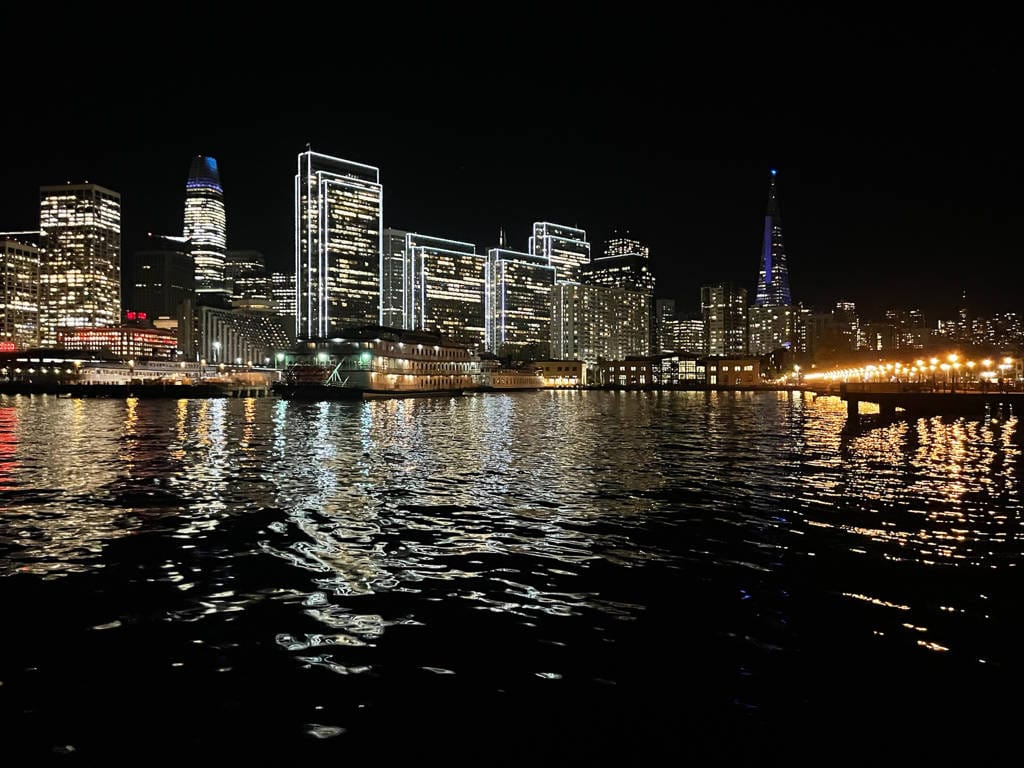 City skyline with illuminated buildings reflected on the water at night; a pier with lights is visible on the right, perfect for Group Events or Private Charters like Alcatraz Island Cruises.