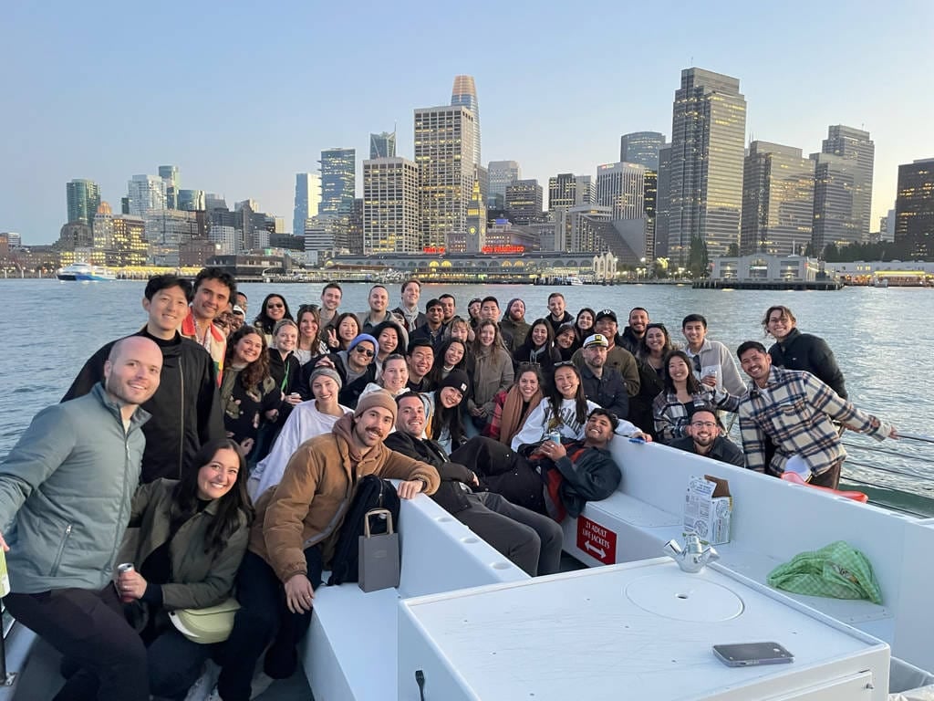 A large group of people pose and smile on a boat with a city skyline and waterfront buildings in the background during daylight, enjoying one of the San Francisco Bay Cruises with Fisherman's Wharf Departures.