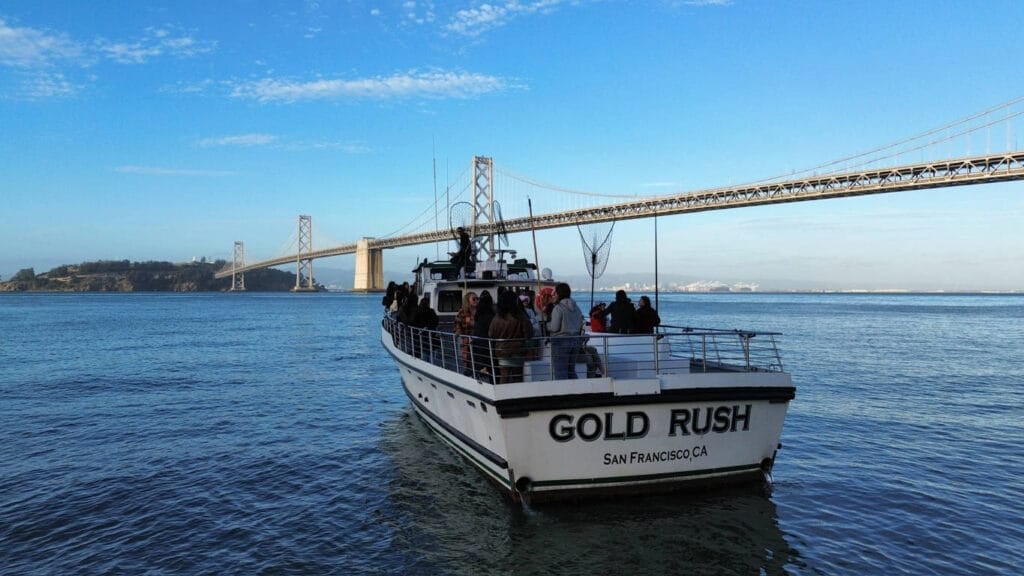 A boat named "Gold Rush" with passengers on board cruises the water near the Bay Bridge in San Francisco, under a clear blue sky—a perfect day for San Francisco Bay Cruises.