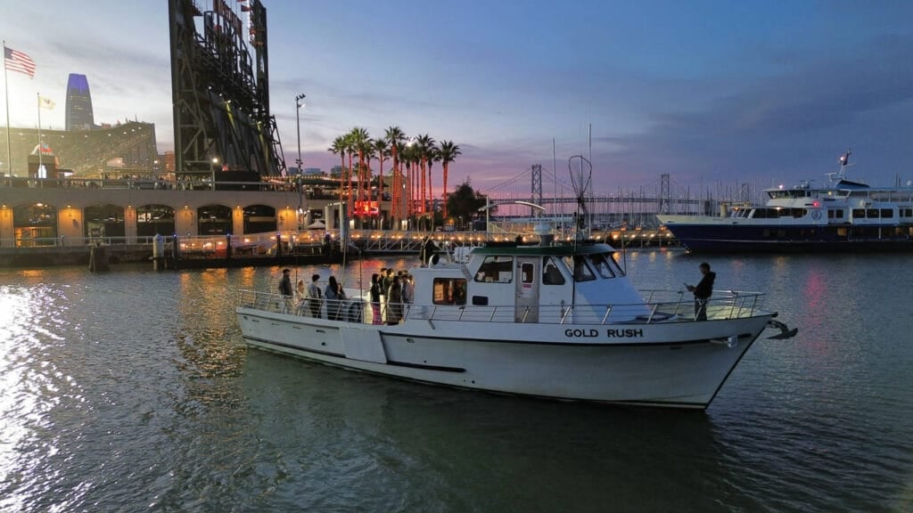 A white boat named "Gold Rush" floats on the water near a marina at dusk, with city lights, palm trees, and a bridge in the background—perfect for San Francisco Bay Cruises or Private Charters departing from Fisherman's Wharf.